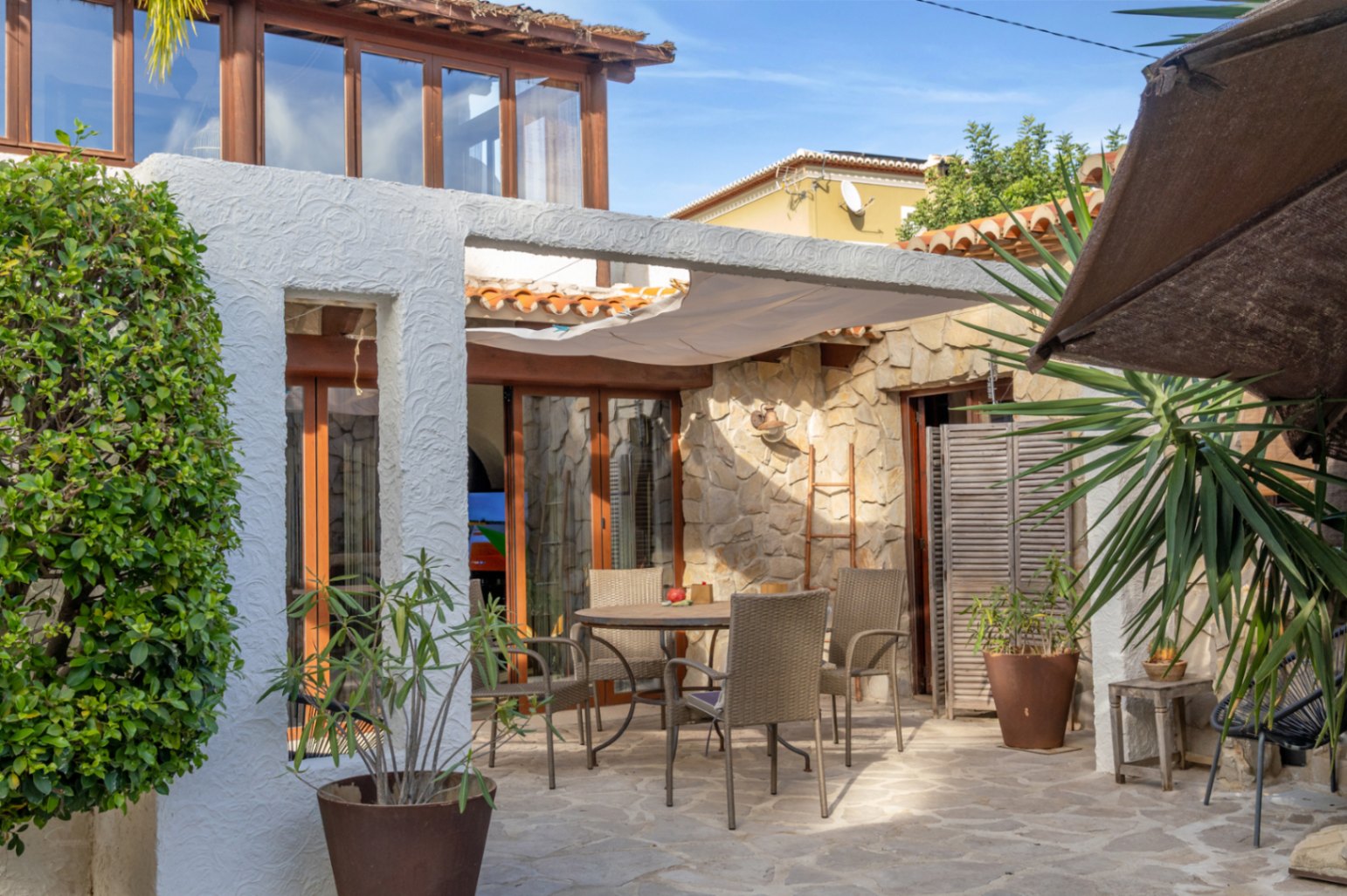 Terraza exterior con suelo de piedra natural, zona de comedor protegida por un toldo vela y fachada que combina mampostería con carpintería de madera.