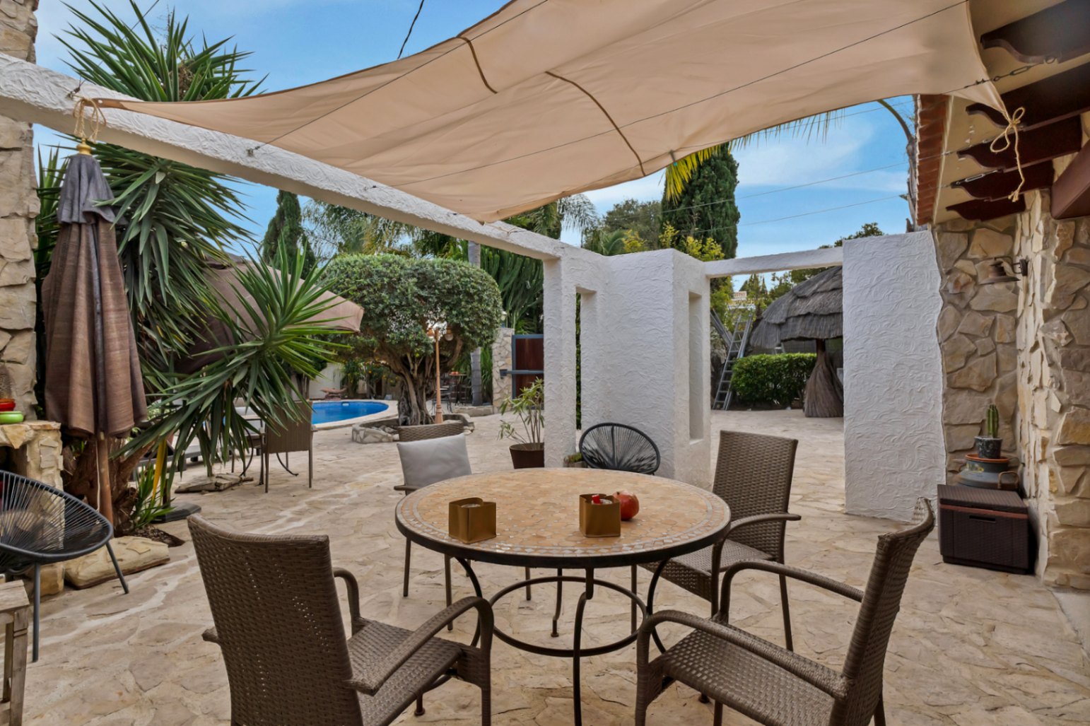 Terraza exterior con suelo de piedra natural, zona de comedor protegida por un toldo vela y vistas al jardín mediterráneo con piscina al fondo.