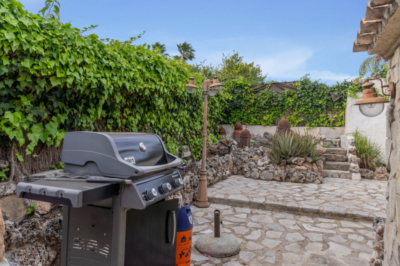 Zona de barbacoa exterior con parrilla de gas Weber, pavimentada con piedra natural y rodeada de muros cubiertos de hiedra y jardinería mediterránea.