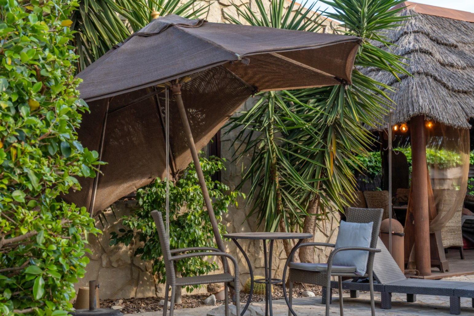Zona de estar exterior con sombrilla, mesa de forja y sillas de mimbre junto a un muro de piedra natural y una pérgola de estilo balinés con techo de paja.