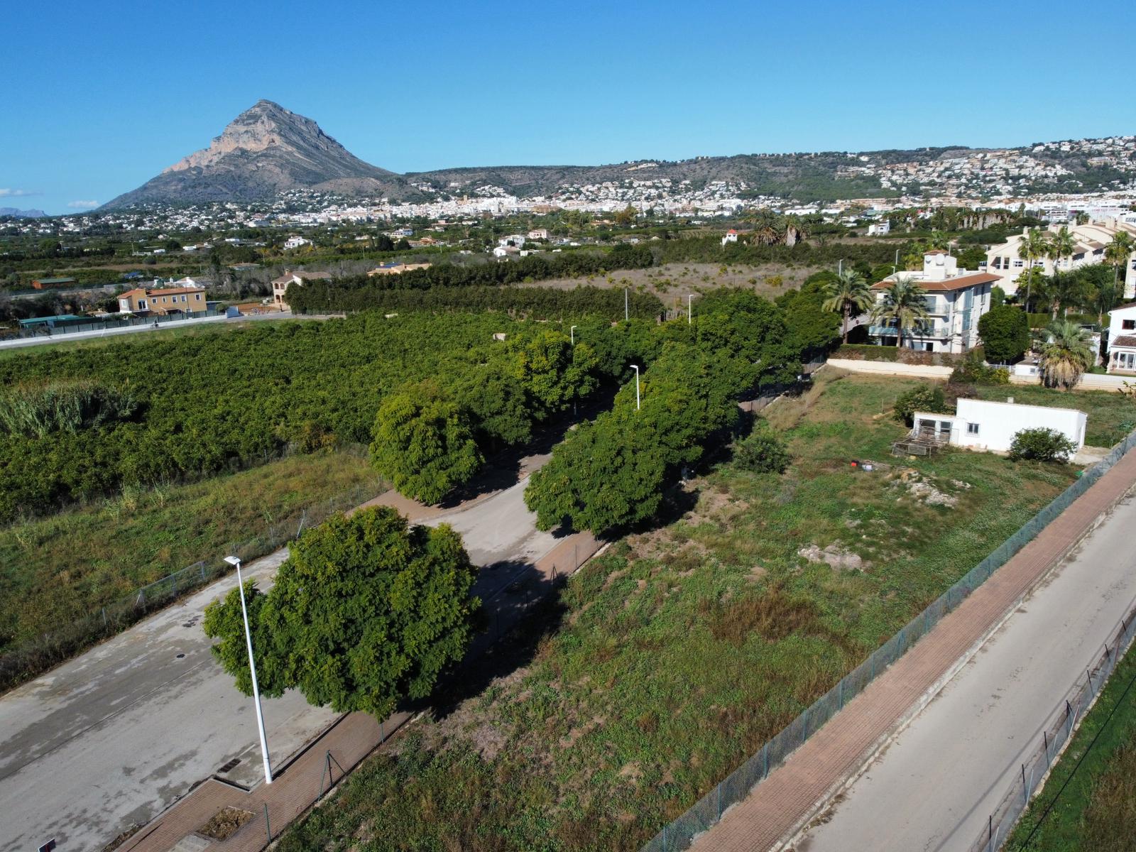 Vista aérea de la parcela residencial en Jávea, mostrando el acceso urbanizado con arbolado y el perfil del Montgó al fondo bajo un cielo despejado.