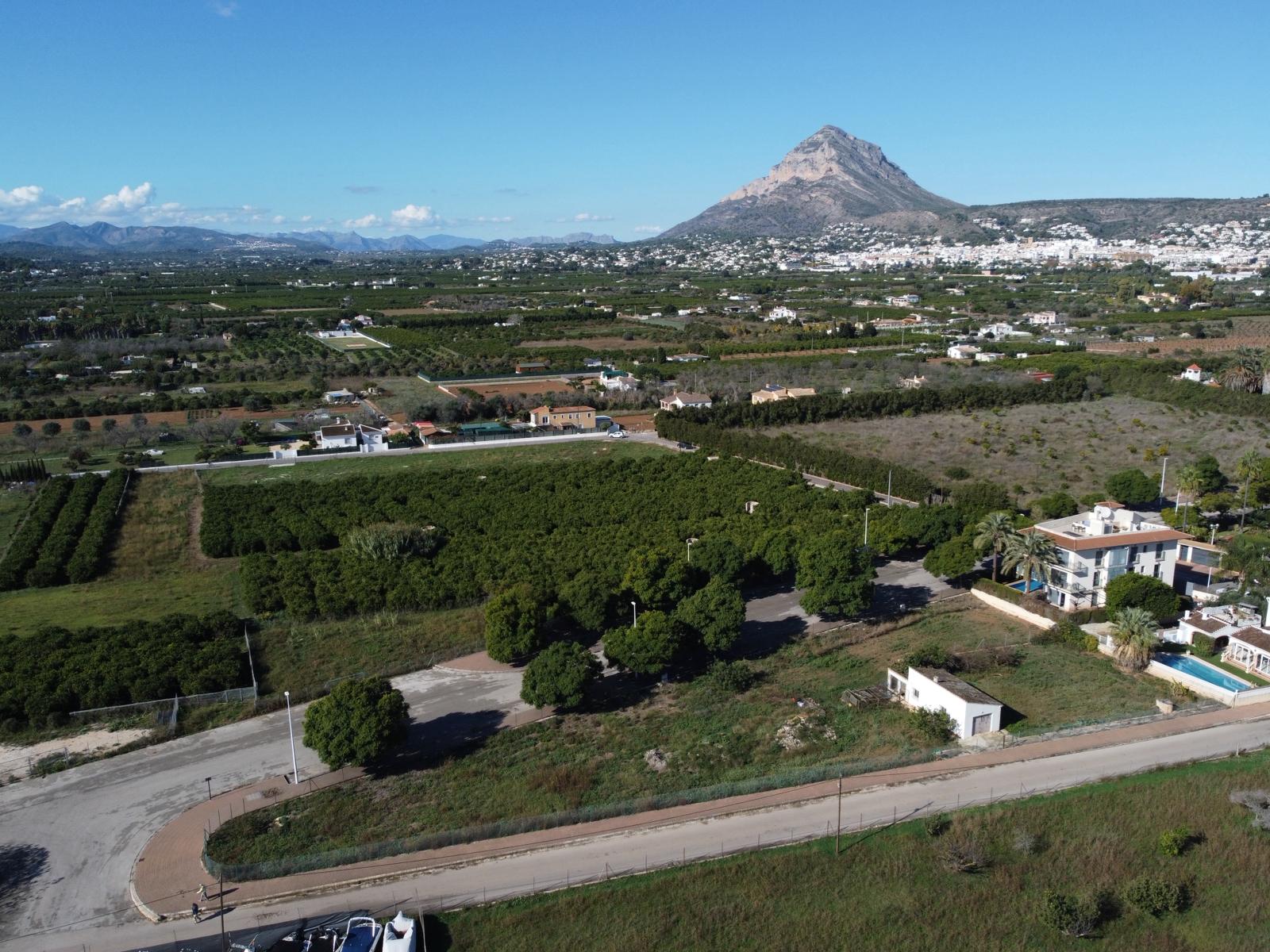 Vista aérea de la parcela urbana con acceso pavimentado, rodeada de zonas verdes y con el imponente macizo del Montgó como telón de fondo.