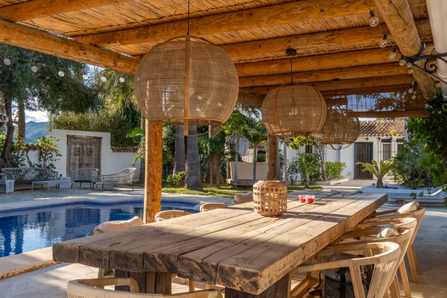 Comedor exterior bajo pérgola de vigas de madera y cañizo, con mesa rústica, lámparas de fibras naturales y vistas directas a la piscina y jardines mediterráneos.