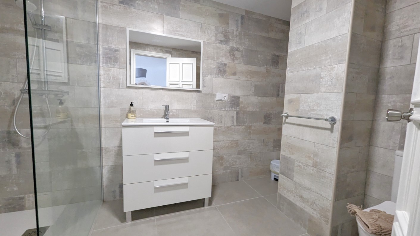 Baño moderno con mueble lavabo blanco de tres cajones, ducha a ras de suelo con mampara de cristal y paredes revestidas en cerámica texturizada de tonos neutros.