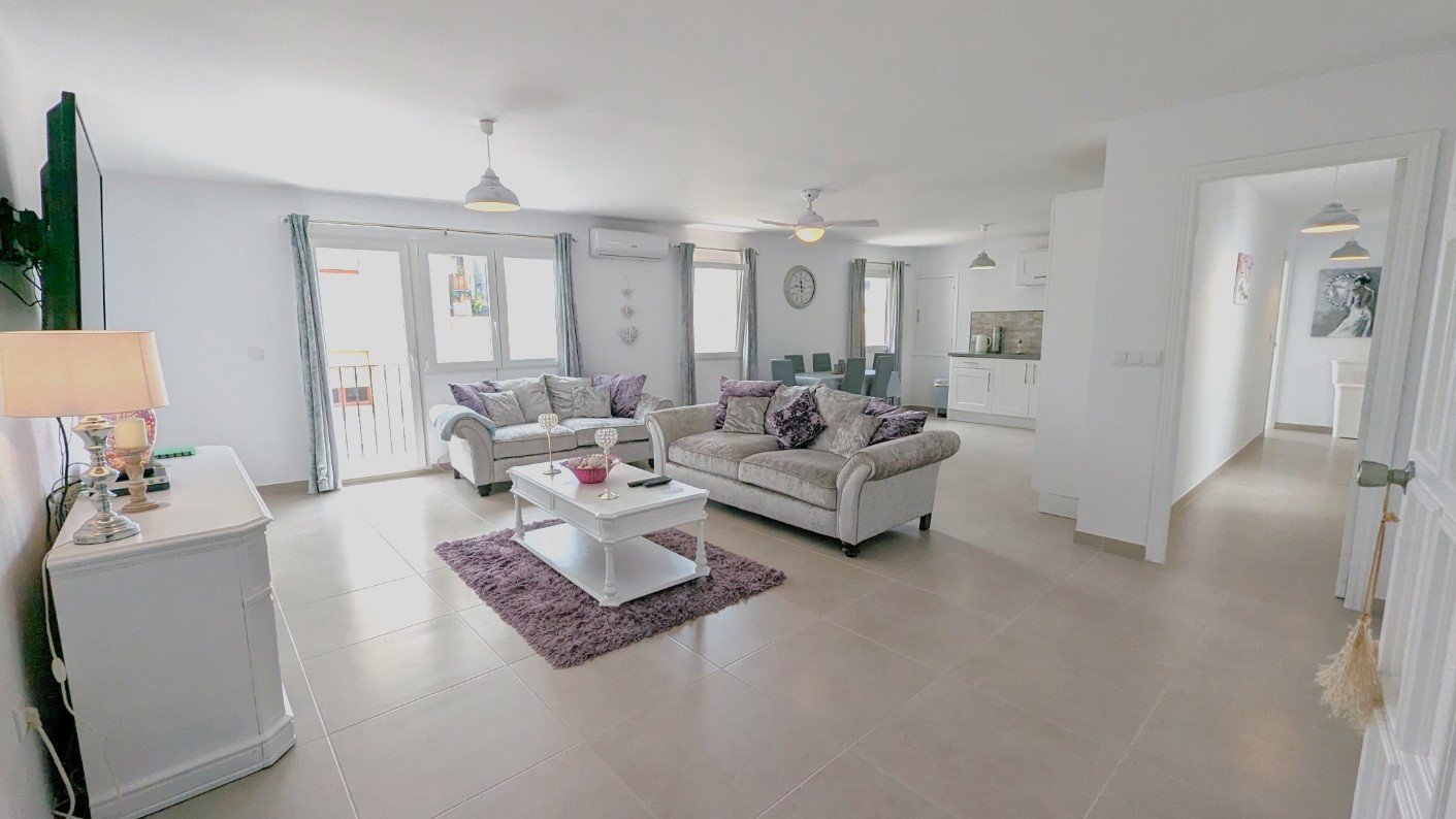 Salón-comedor de planta abierta con sofás grises, detalles en violeta y cocina blanca integrada. Dispone de suelos cerámicos, aire acondicionado y acceso a balcones exteriores.