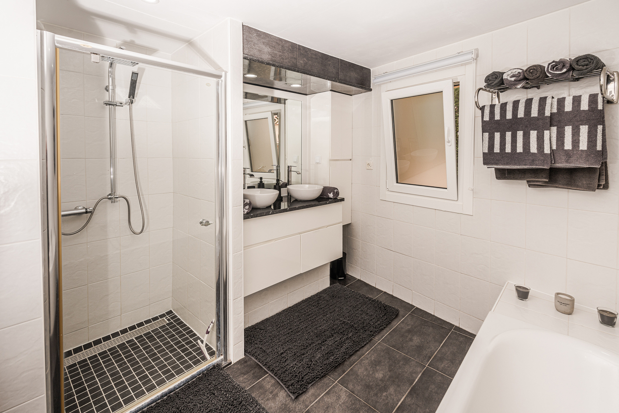 Baño moderno con ducha de cristal, lavabo doble sobre encimera oscura y suelos de gres porcelánico en contraste con azulejos blancos.