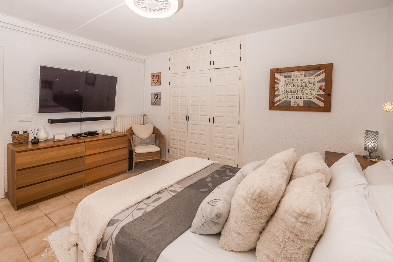 Dormitorio con armarios empotrados blancos, televisor de gran formato sobre cómoda de madera y suelos de gres. Incluye calefacción por radiador y un sillón de mimbre.