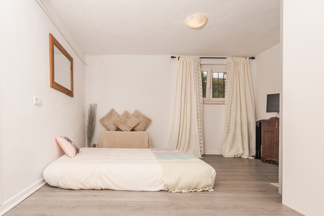 Dormitorio con suelo laminado gris, paredes blancas y cama de perfil bajo. Incluye un mueble de madera oscura, espejo con marco de madera y ventana con cortinas neutras.
