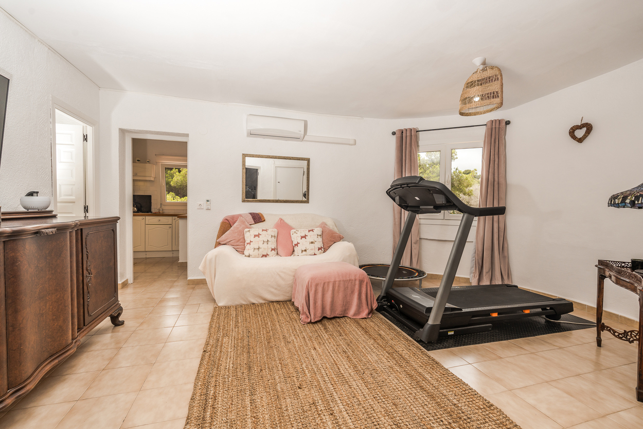 Sala de estar polivalente con zona de fitness, cinta de correr, suelos cerámicos y aire acondicionado, que conecta visualmente con la cocina.