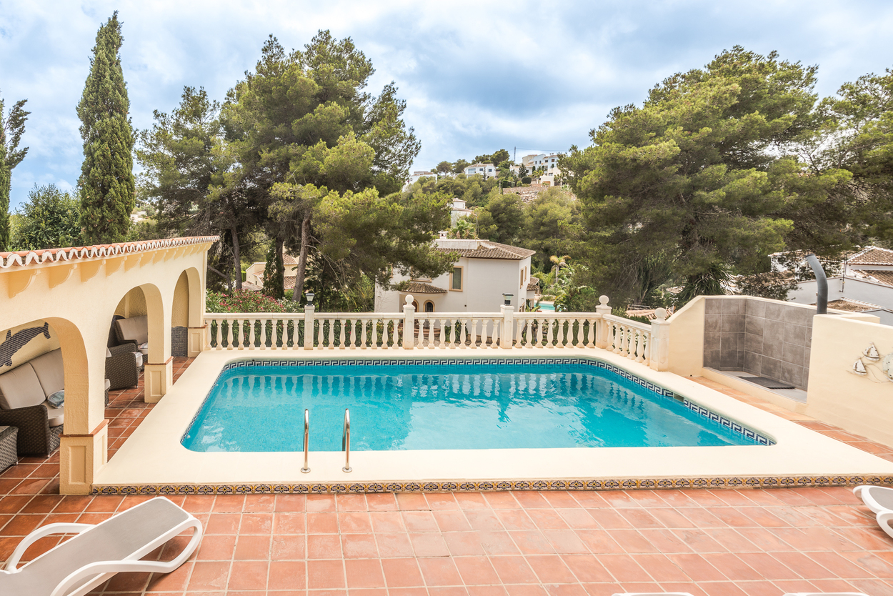 Terraza exterior con piscina de 8x4m, balaustrada blanca y una naya cubierta con arcos y zona de estar, rodeada de pinos maduros y vegetación mediterránea.