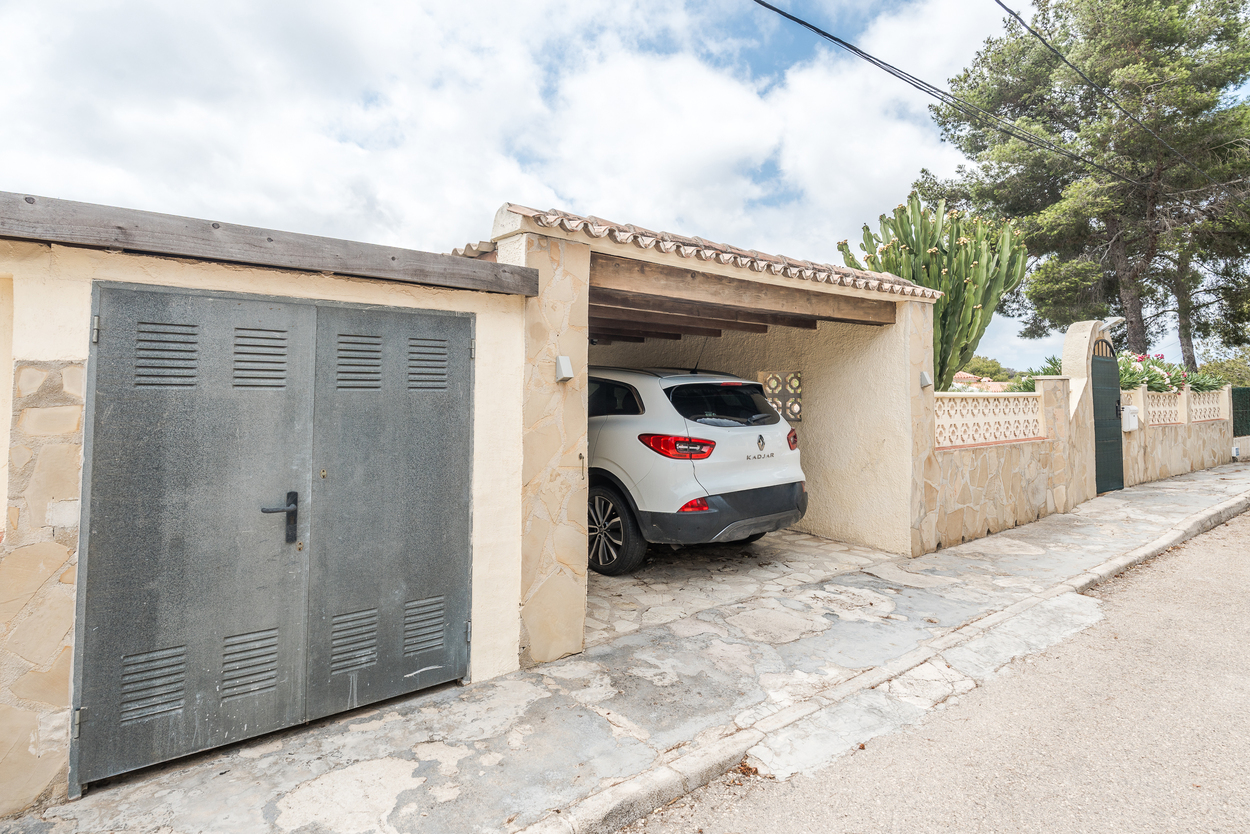 Vista exterior que muestra la entrada vallada, muros revestidos de piedra y un porche cubierto con vigas de madera para aparcamiento seguro.