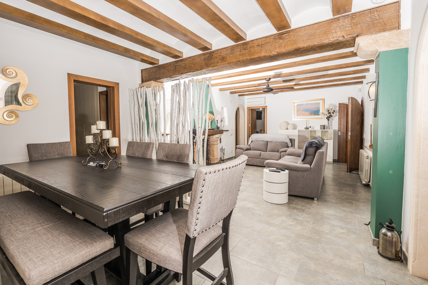 Amplio salón-comedor con vigas de madera a la vista, suelos de piedra y una mesa de madera oscura con sillas tapizadas en tonos grises.