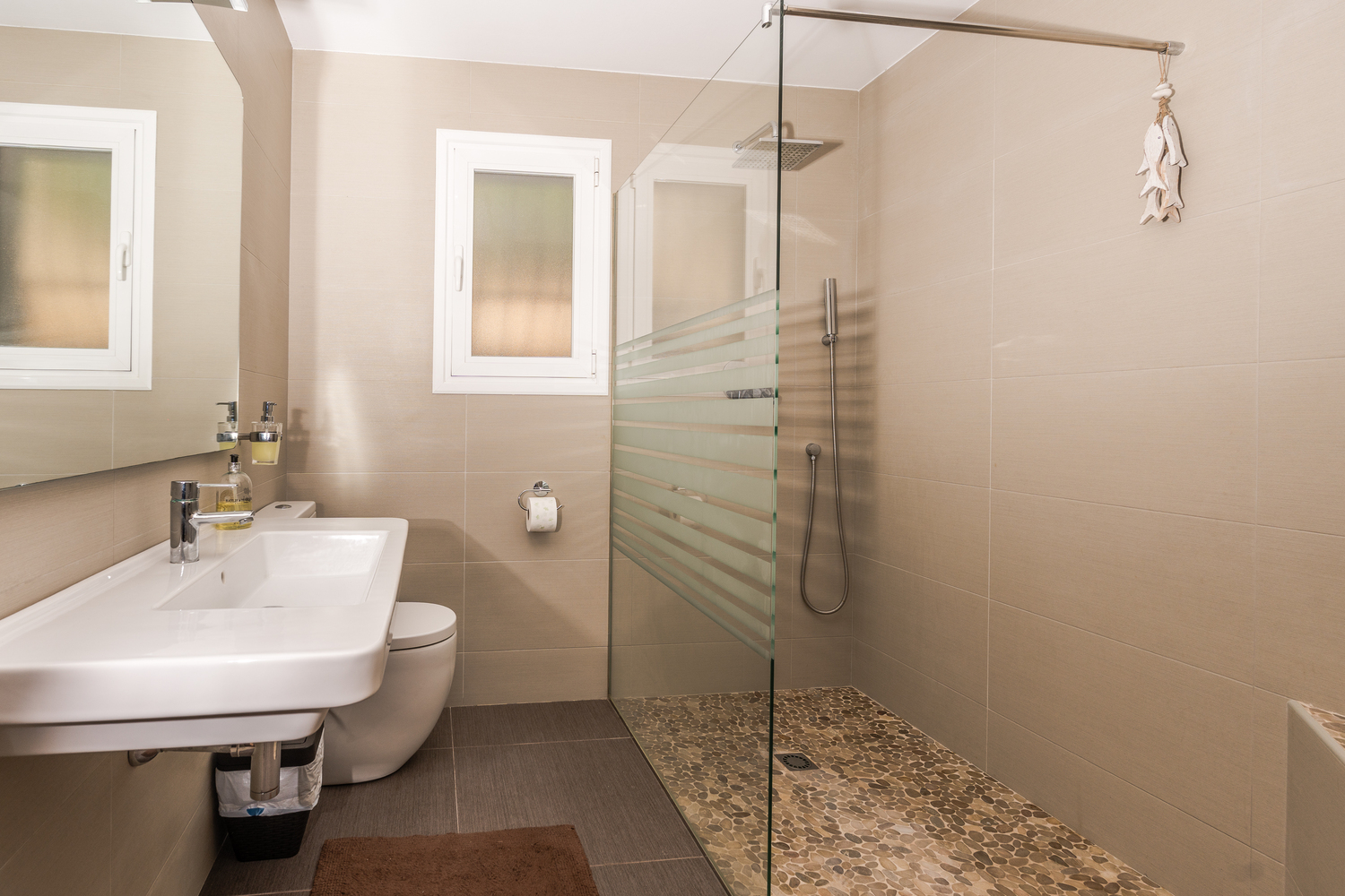 Baño moderno con ducha a ras de suelo, suelo de cantos rodados, mampara de cristal y azulejos en tonos neutros. Incluye lavabo suspendido y ventana translúcida.