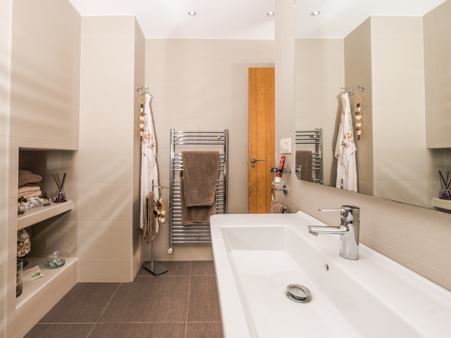Baño moderno con lavabo cerámico de gran formato, estanterías de obra integradas y radiador toallero cromado sobre revestimiento en tonos beige.