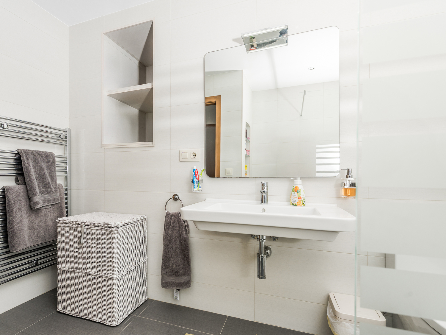 Baño moderno con lavabo suspendido, hornacina de pared integrada y radiador toallero cromado sobre azulejos blancos y suelo gris oscuro.