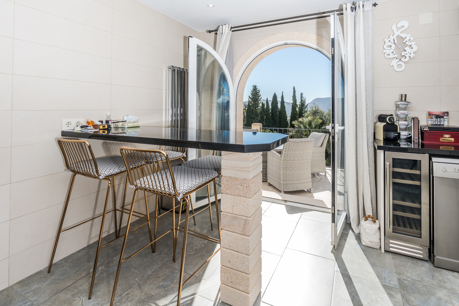 Barra de cocina con encimera de granito negro y taburetes dorados, conectada a una terraza exterior mediante un arco de piedra con vistas al valle y las montañas.