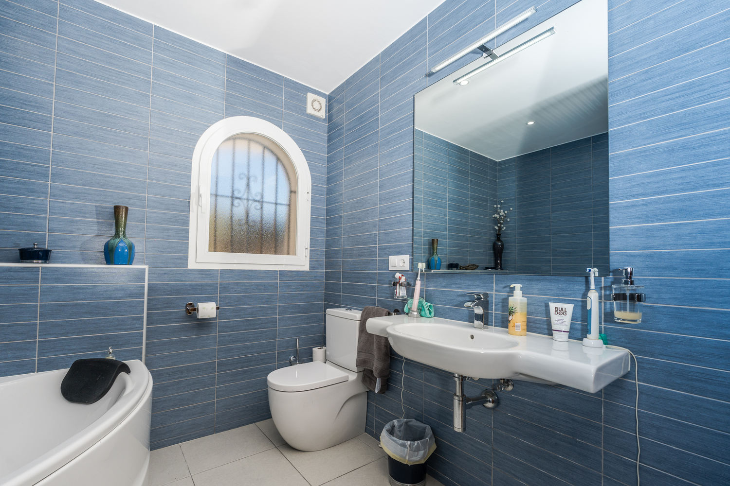 Cuarto de baño moderno con azulejos azules de textura horizontal, ventana de arco con cristal translúcido, lavabo blanco y un gran espejo con iluminación superior.