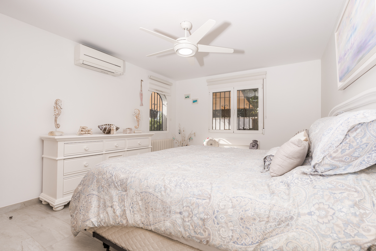Dormitorio luminoso con paredes blancas, ventilador de techo, aire acondicionado y cómoda blanca. Incluye ventanas con rejas de seguridad y suelos cerámicos.