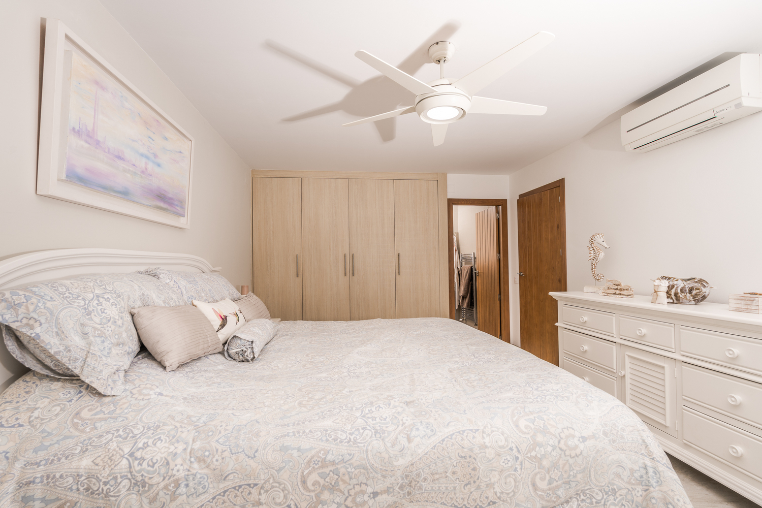 Dormitorio moderno con armarios empotrados de madera clara, cómoda blanca y ventilador de techo. Incluye aire acondicionado y acceso directo a una estancia contigua.