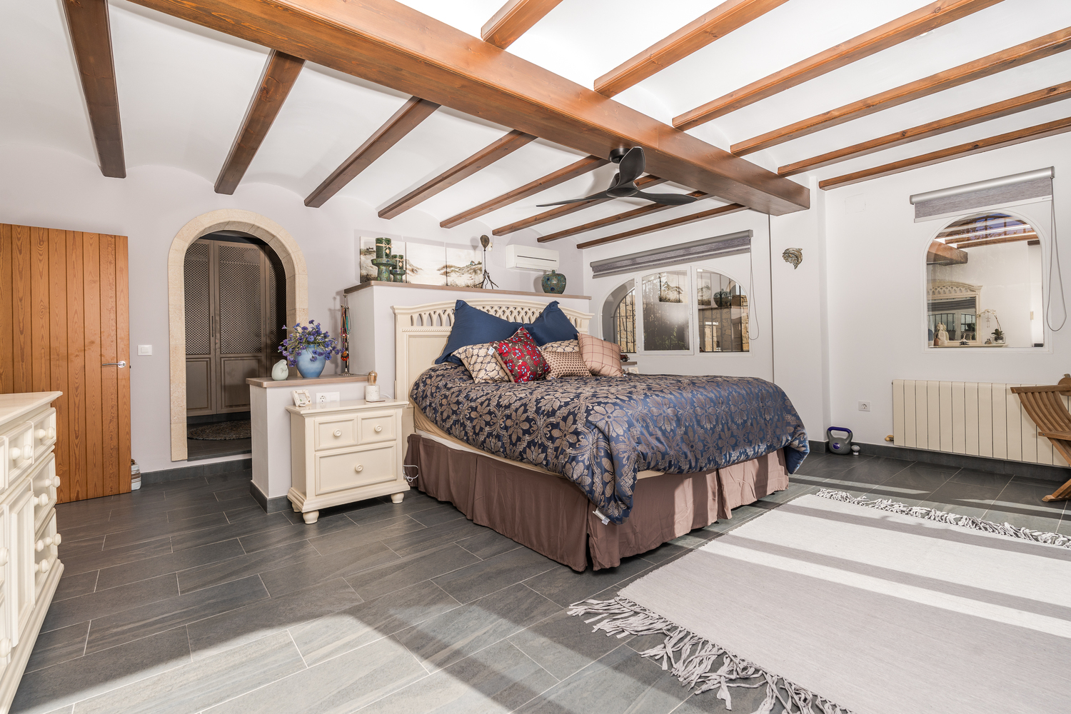 Dormitorio principal con vigas de madera vista, suelo de baldosas grises y un arco de piedra natural que da acceso al vestidor. Equipado con aire acondicionado y calefacción.