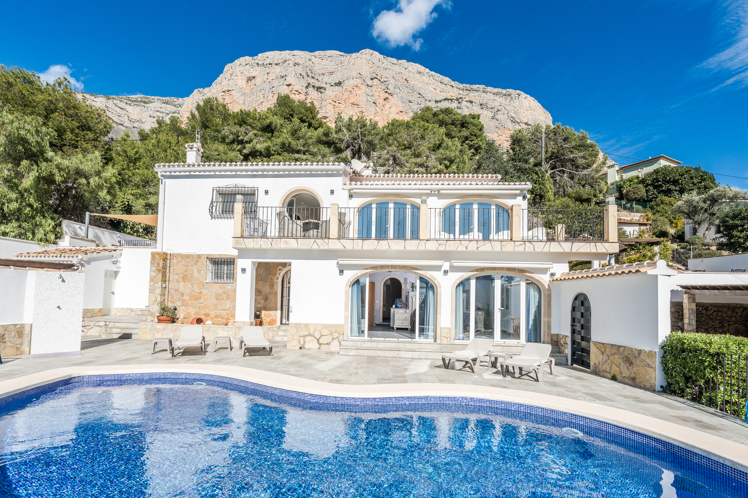 Fachada de la villa con piscina curva, terraza de piedra y ventanales en arco, destacando la imponente pared rocosa del Montgó al fondo.
