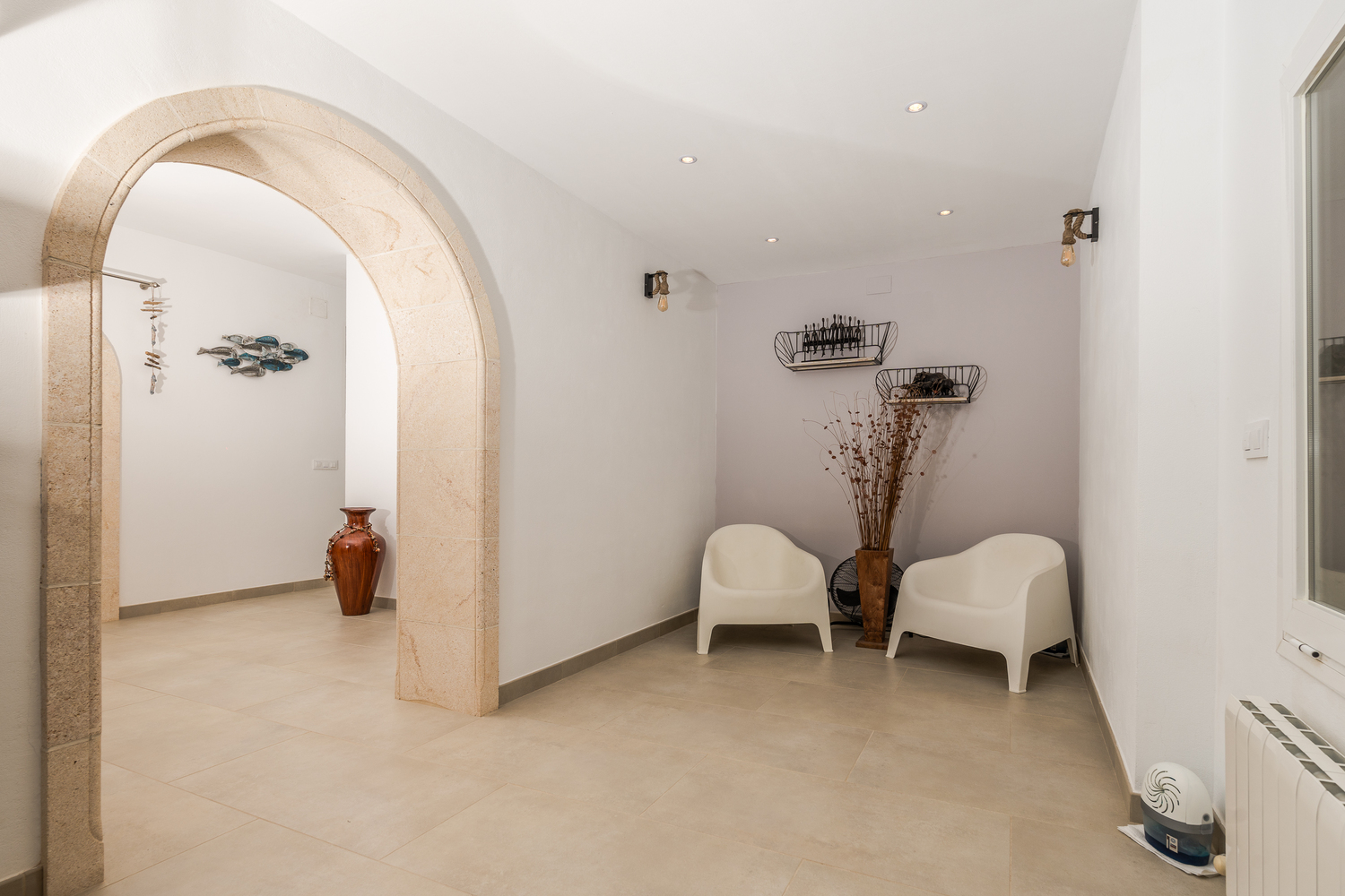 Pasillo con un arco tradicional de piedra Tosca, iluminación empotrada y una zona de estar minimalista con sillones blancos y apliques de pared de estilo industrial.