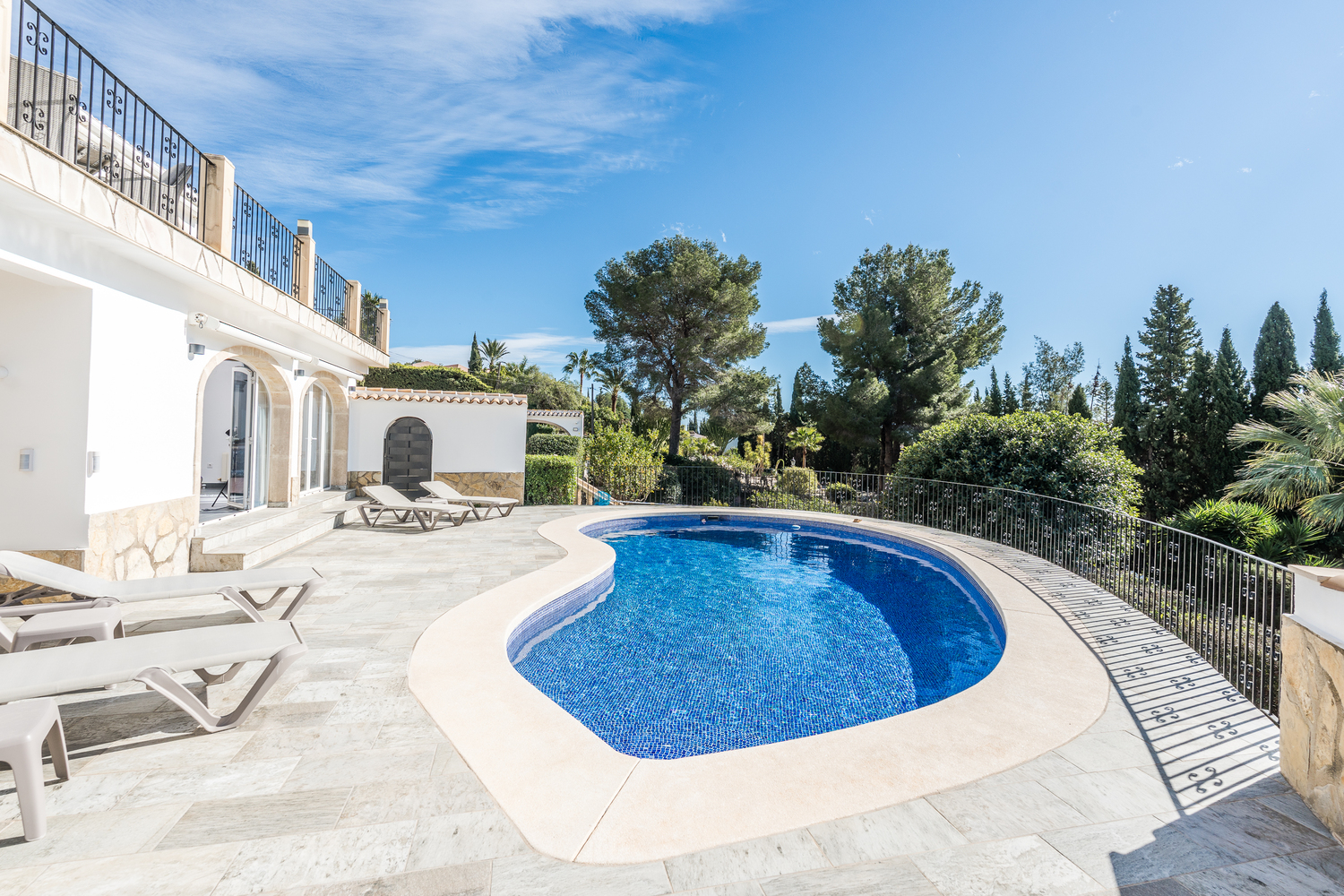 Piscina de forma arriñonada rodeada por una terraza de piedra clara con tumbonas, enmarcada por vegetación mediterránea y la fachada blanca de la villa.