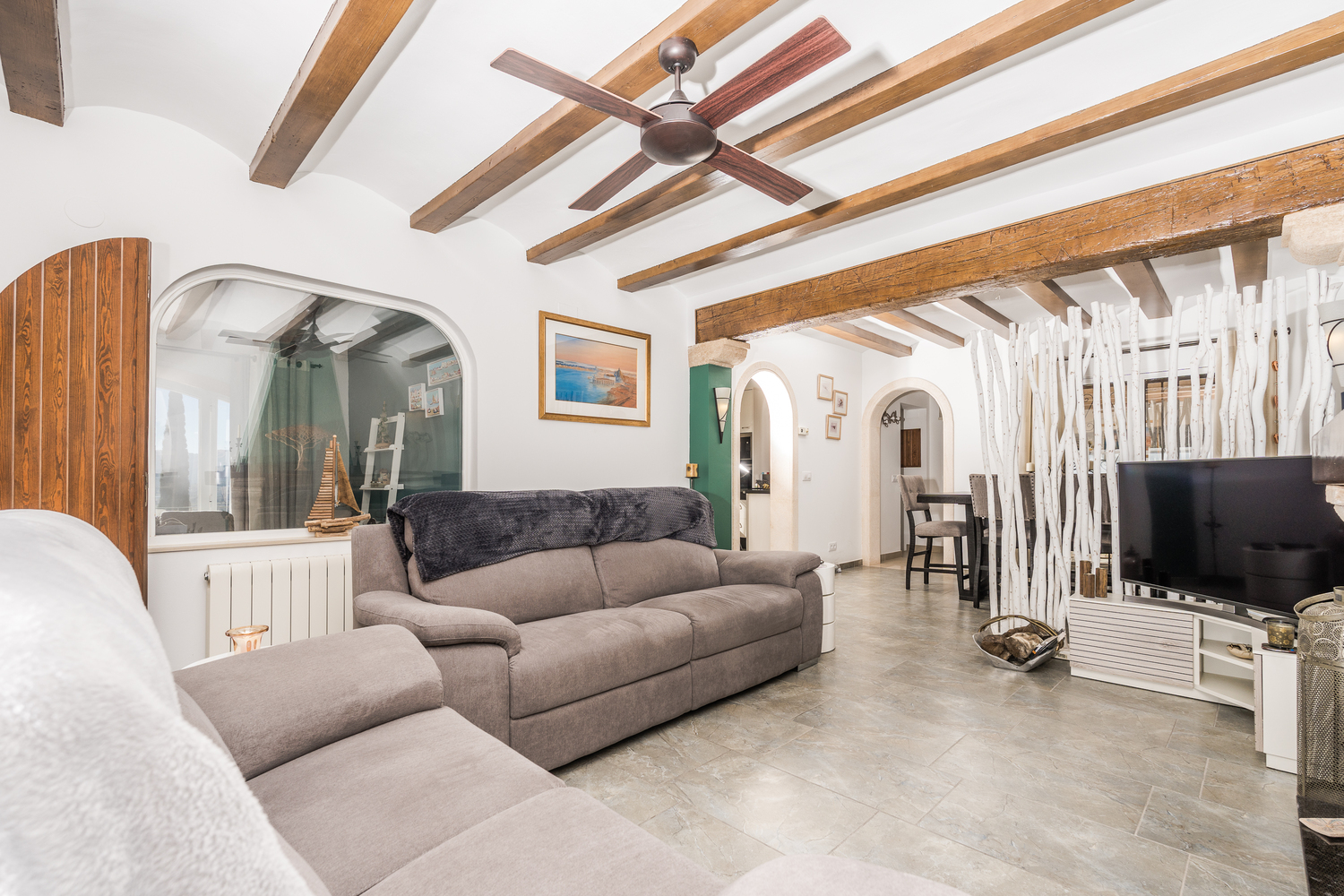 Salón con vigas de madera vistas, ventilador de techo y suelos cerámicos. Un separador decorativo de troncos blancos divide la zona de estar del comedor al fondo.