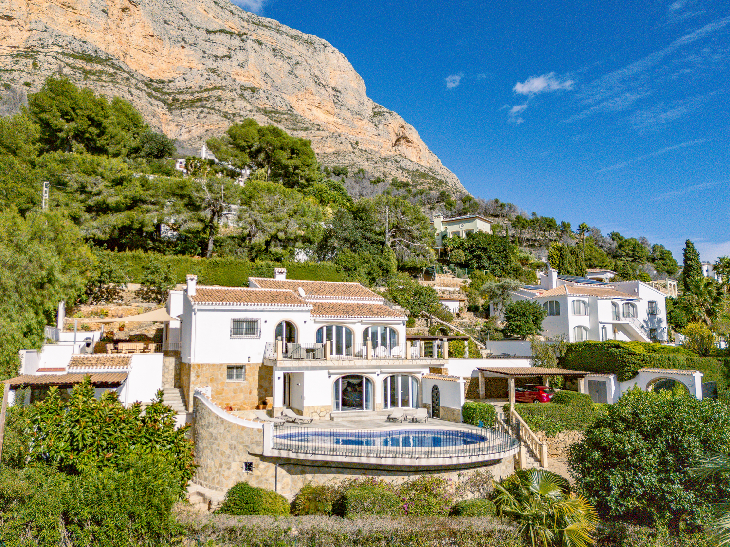 Vista exterior de la villa de dos niveles al pie del Montgó, con piscina curva, fachadas blancas con zócalos de piedra y naya acristalada con arcos.