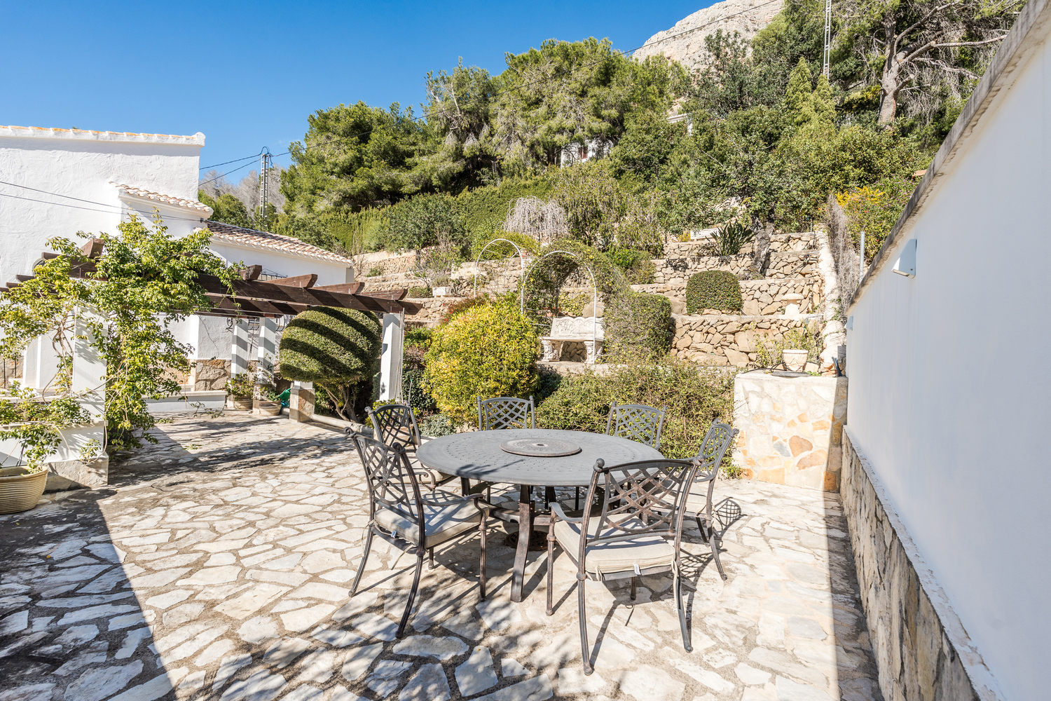Zona de comedor exterior con mesa de hierro forjado y pavimentación de piedra irregular, rodeada de jardines en terrazas y una pérgola de madera.
