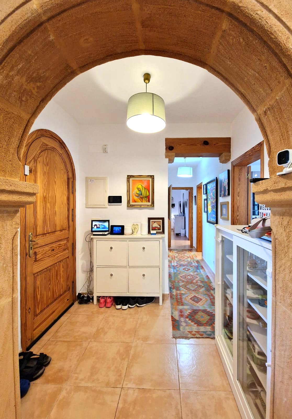 Recibidor visto a través de un arco de piedra Tosca, con una puerta de madera arqueada, suelos cerámicos y un pasillo con alfombra de motivos geométricos y vigas de madera.