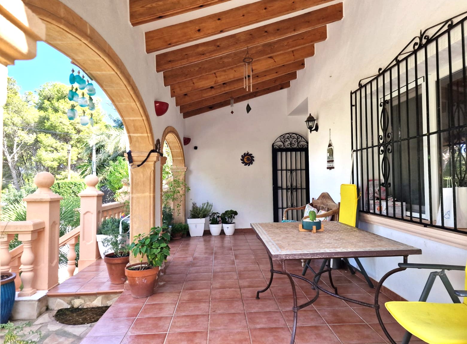 Terraza cubierta con arcos de piedra Tosca, vigas de madera a la vista y suelo de terracota. Incluye zona de comedor exterior con vistas a la vegetación mediterránea.