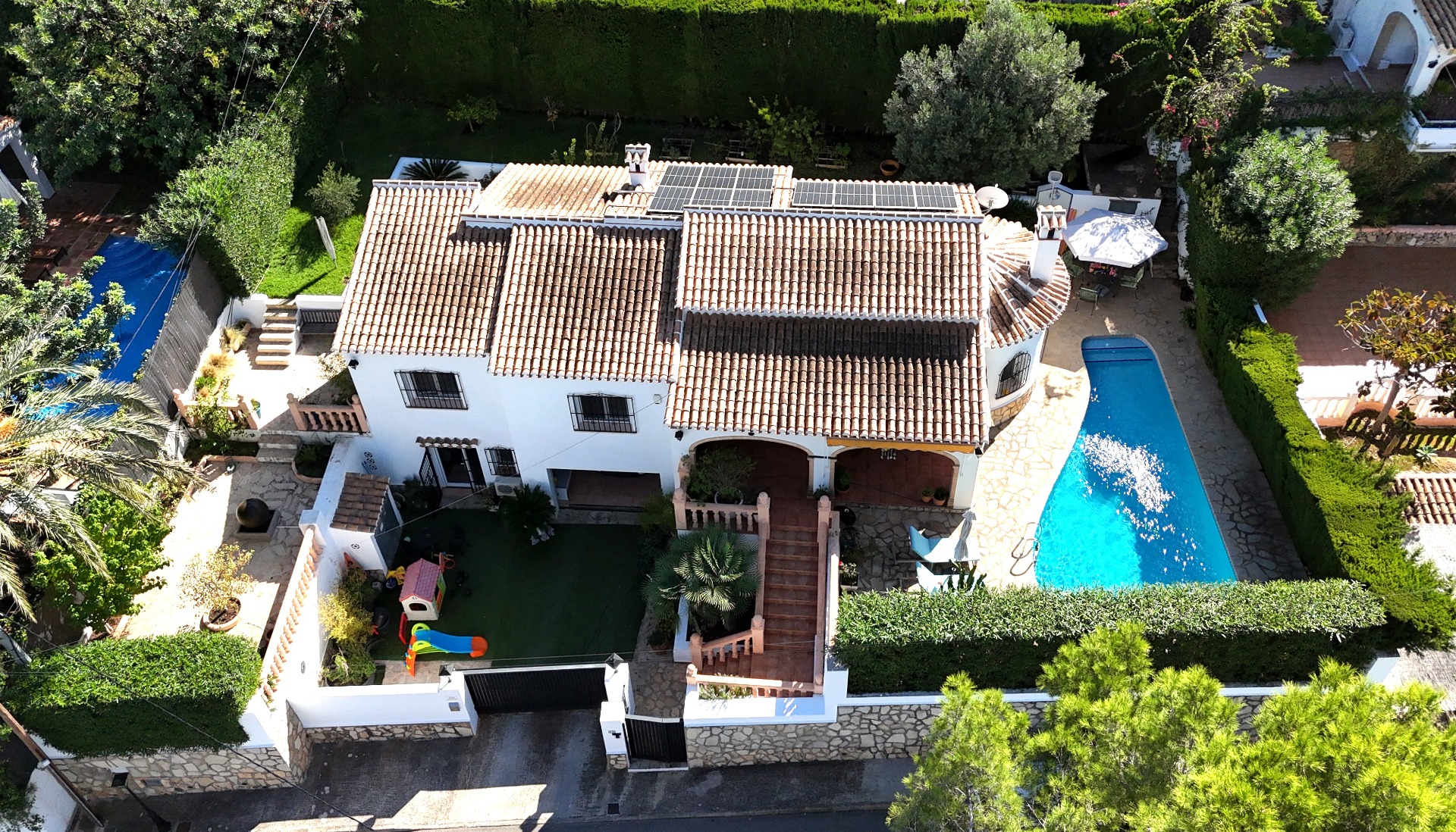 Vista aérea de esta villa en Toscal con tejado de terracota, paneles solares, piscina privada y zona de juegos infantil rodeada de vegetación mediterránea.