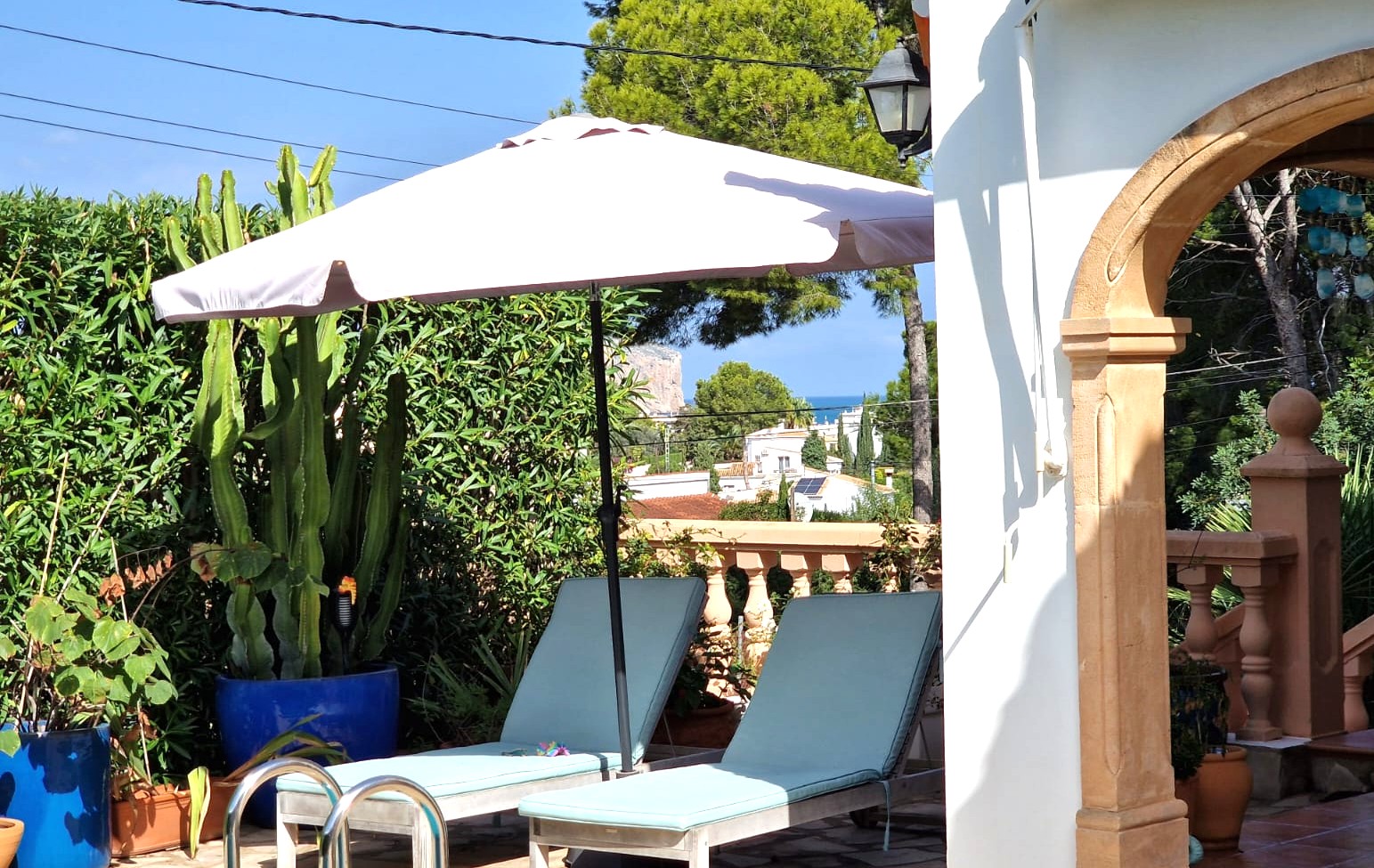 Zona de terraza con tumbonas y sombrilla junto a un arco de piedra tosca, rodeada de vegetación mediterránea y con vistas lejanas al mar y al Cabo de San Antonio.