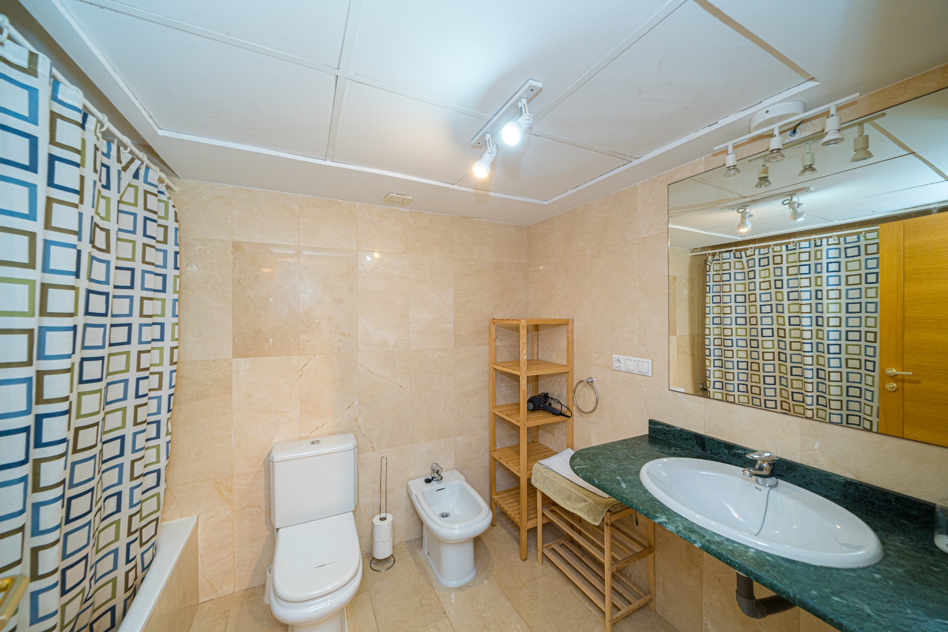 Baño moderno con revestimiento de mármol crema, encimera de mármol verde y sanitarios blancos. Incluye bidé, estanterías de madera y sistema de iluminación por focos.