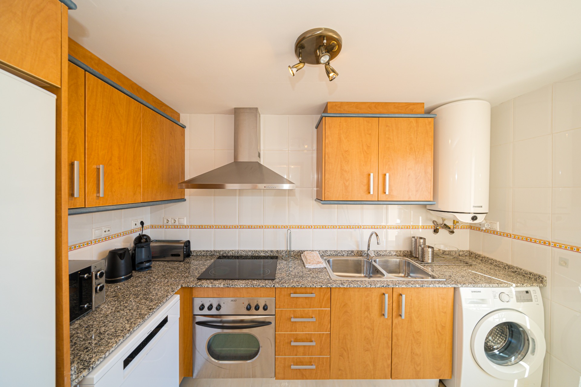 Cocina equipada con armarios de madera, encimera de granito y electrodomésticos integrados, incluyendo horno, lavadora y campana extractora de acero inoxidable.