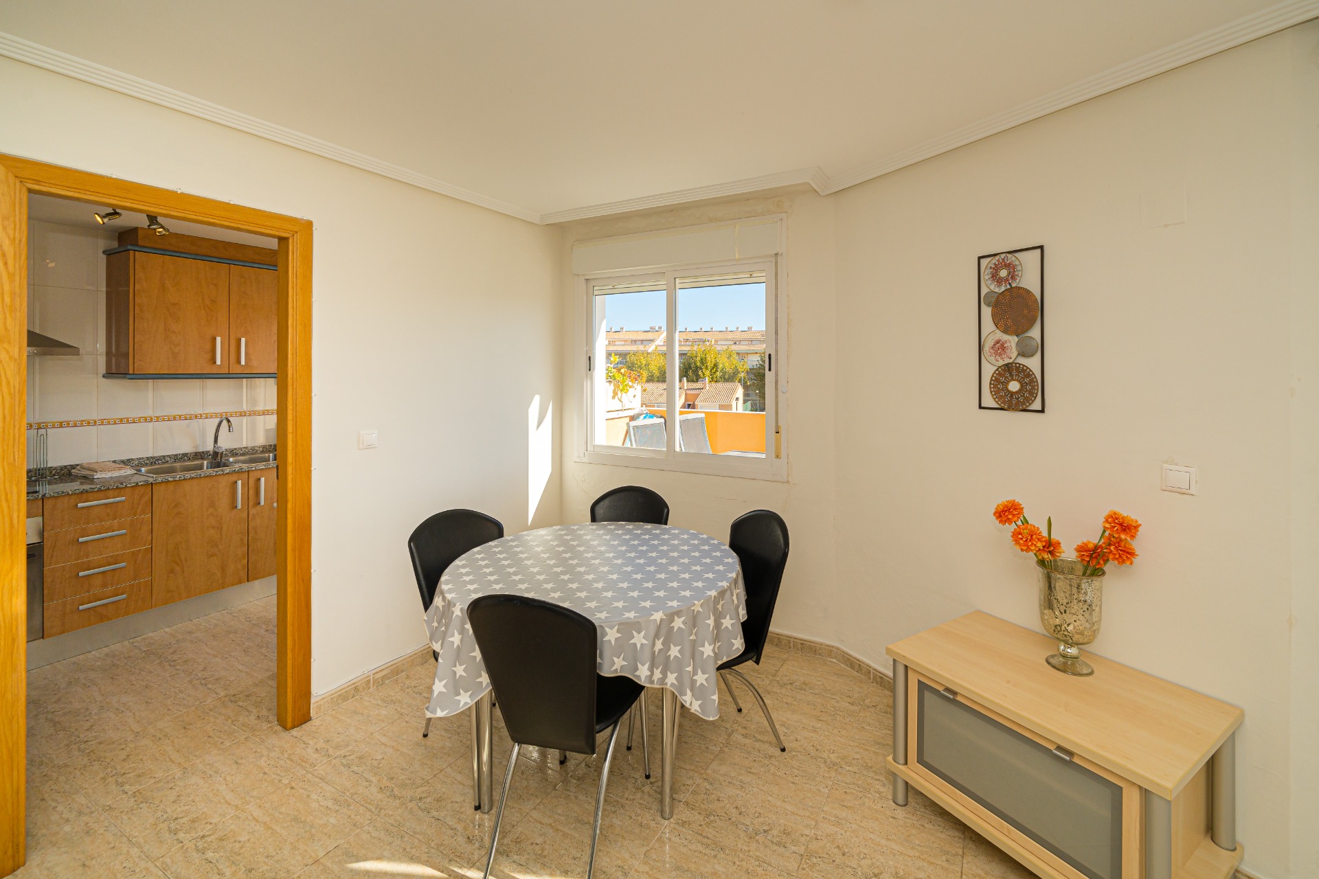 Comedor luminoso con mesa redonda y suelos de gres, situado junto a la cocina de madera. El ventanal aporta luz natural y vistas despejadas al entorno urbano.