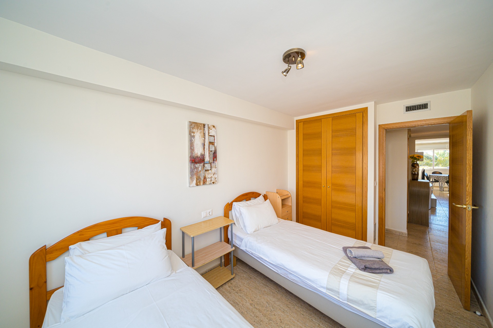 Dormitorio luminoso con dos camas individuales, cabeceros de madera, armario empotrado de roble y suelos de gres. La puerta abierta conecta con el resto de la vivienda.