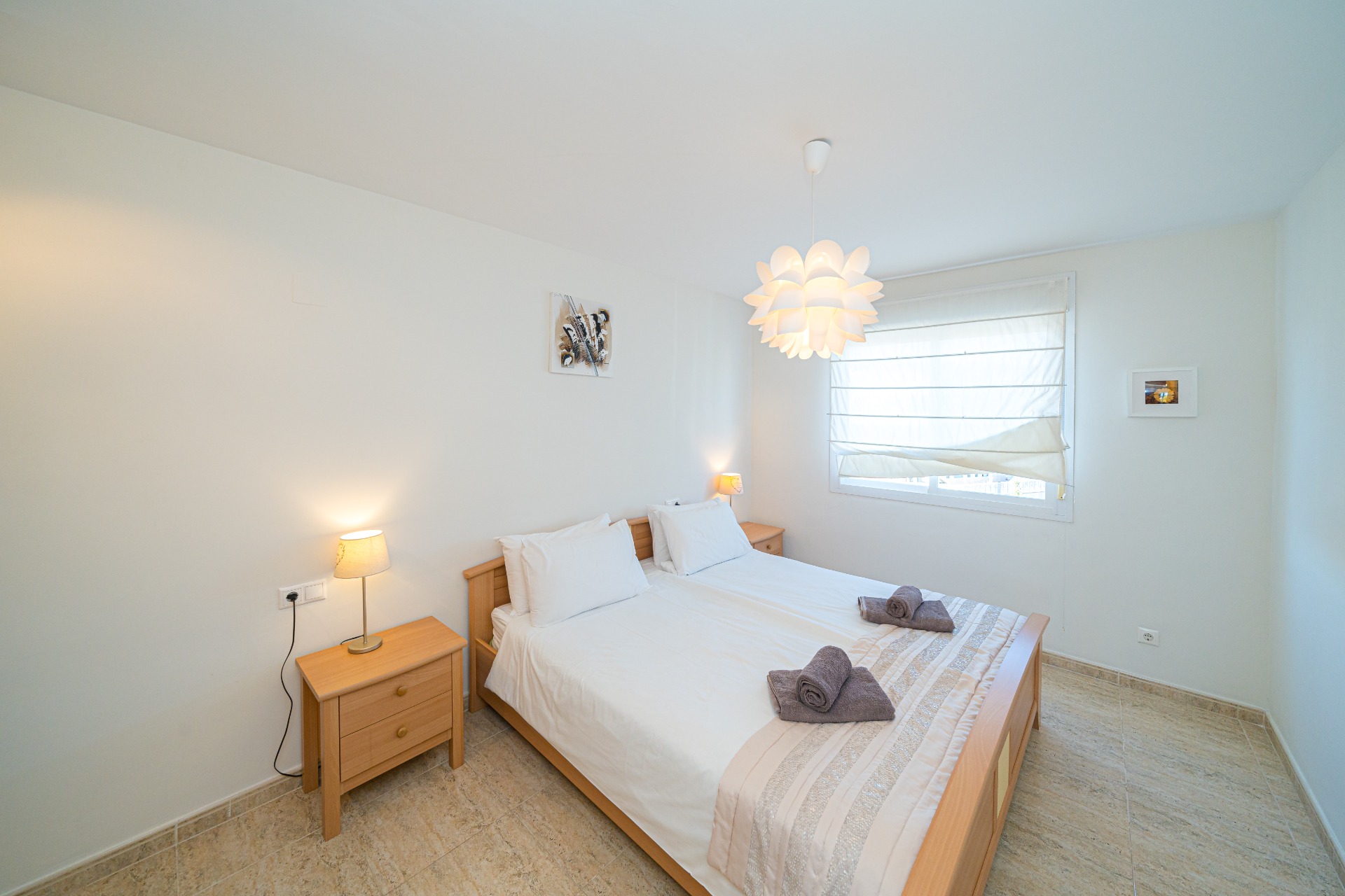 Dormitorio luminoso con mobiliario de madera clara, cama doble y mesitas de noche a juego. La luz natural entra por la ventana con estor blanco sobre suelos cerámicos.