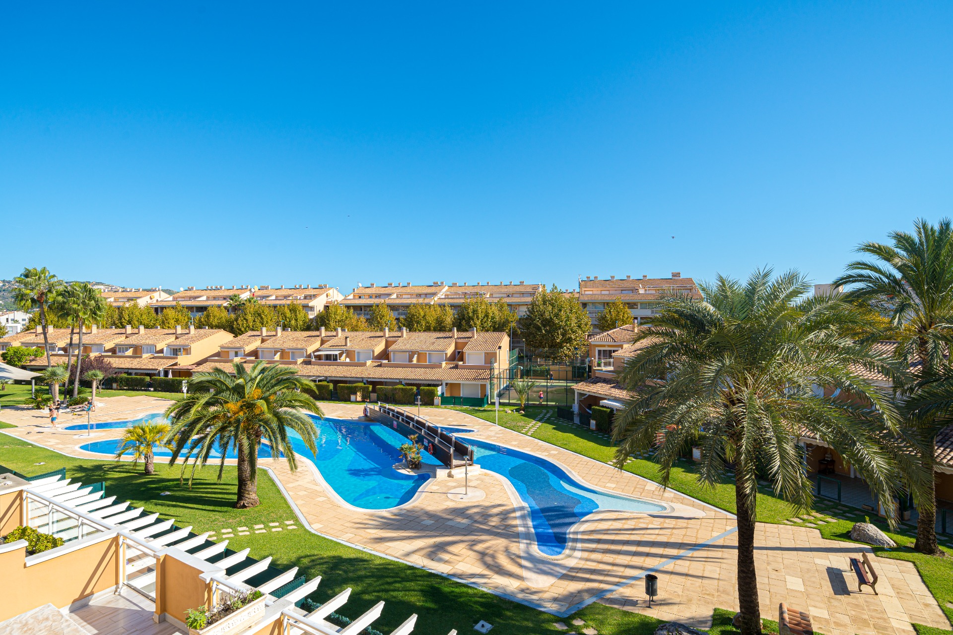 Vista de la amplia piscina comunitaria de estilo laguna con puente central, rodeada de palmeras y jardines cuidados en el complejo Moreras del Saladar.