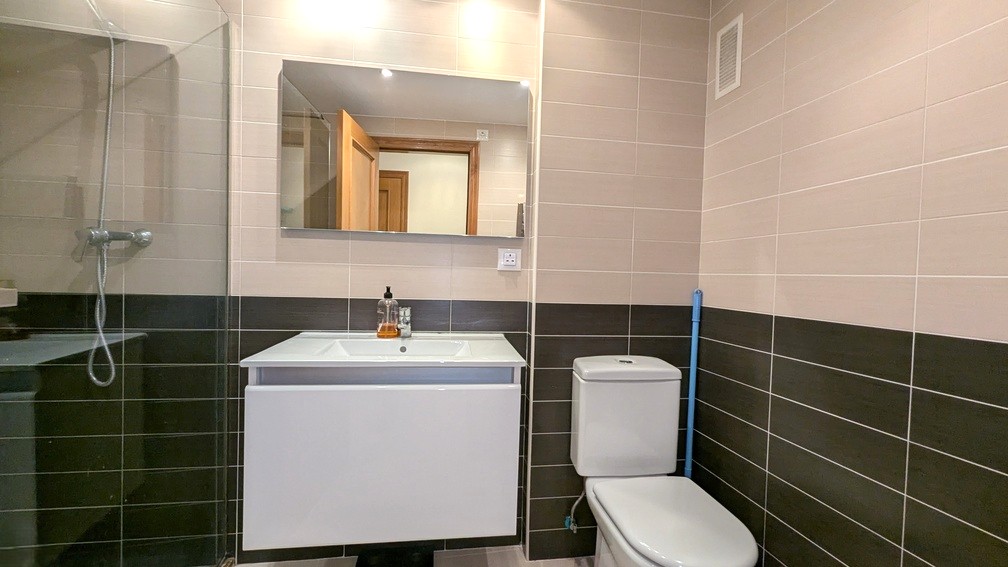 Baño moderno con azulejos bicolores, lavabo suspendido blanco, espejo rectangular y zona de ducha con mampara de cristal fija.