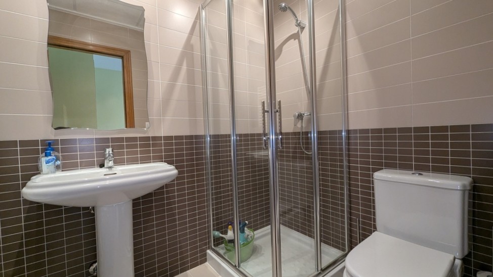 Cuarto de baño funcional con ducha de esquina acristalada, lavabo de pedestal y azulejos en tonos marrón y crema.
