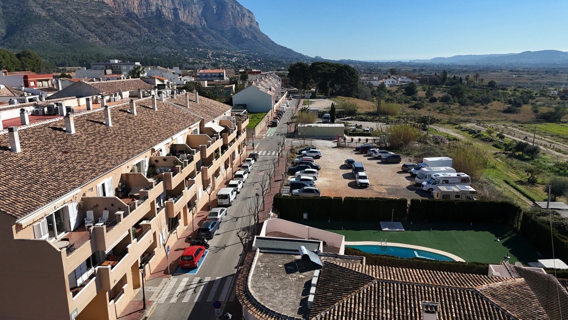 Vista elevada del complejo residencial que incluye la piscina comunitaria, el aparcamiento y vistas panorámicas al Montgó y al paisaje rural circundante.