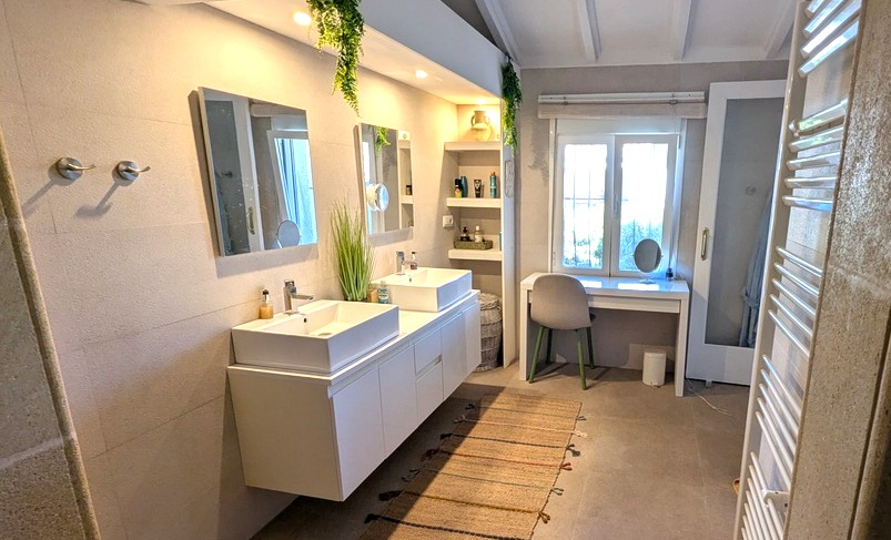 Baño moderno con lavabo de doble seno, mueble suspendido y estanterías integradas. Dispone de un tocador bajo la ventana y techos con vigas de madera blancas.