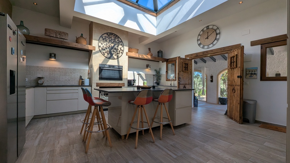 Cocina moderna con una cúpula de cristal central, isla con taburetes y puertas de madera tradicionales que conectan directamente con la naya exterior.