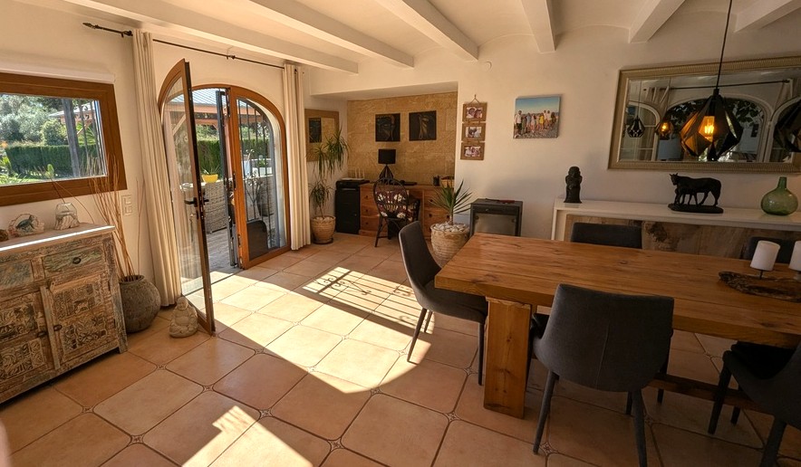Comedor de planta abierta con vigas blancas vistas, suelos de terracota y mesa de madera maciza. Una puerta de arco acristalada conecta directamente con la terraza exterior.