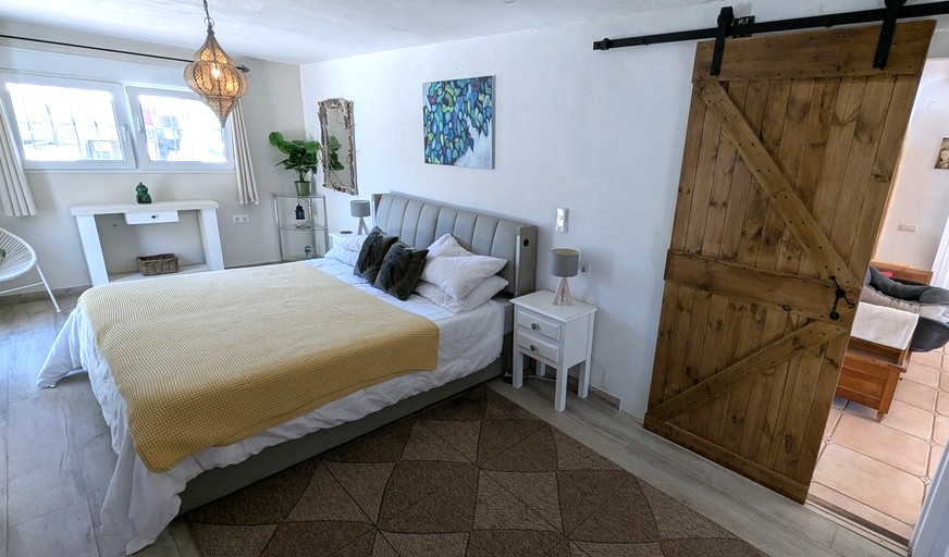 Dormitorio luminoso con puerta corredera de madera estilo granero, cabecero tapizado y lámpara colgante de estilo marroquí sobre suelo cerámico imitación madera.
