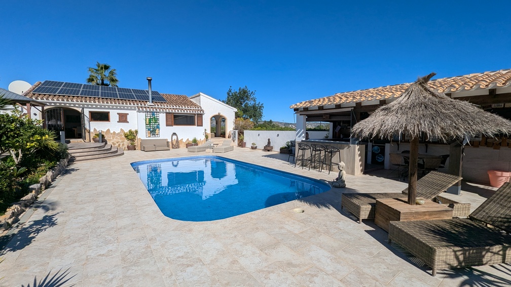 Exterior de la villa principal con piscina privada, terraza de piedra y cocina de verano con barra, destacando los paneles solares instalados en el tejado.