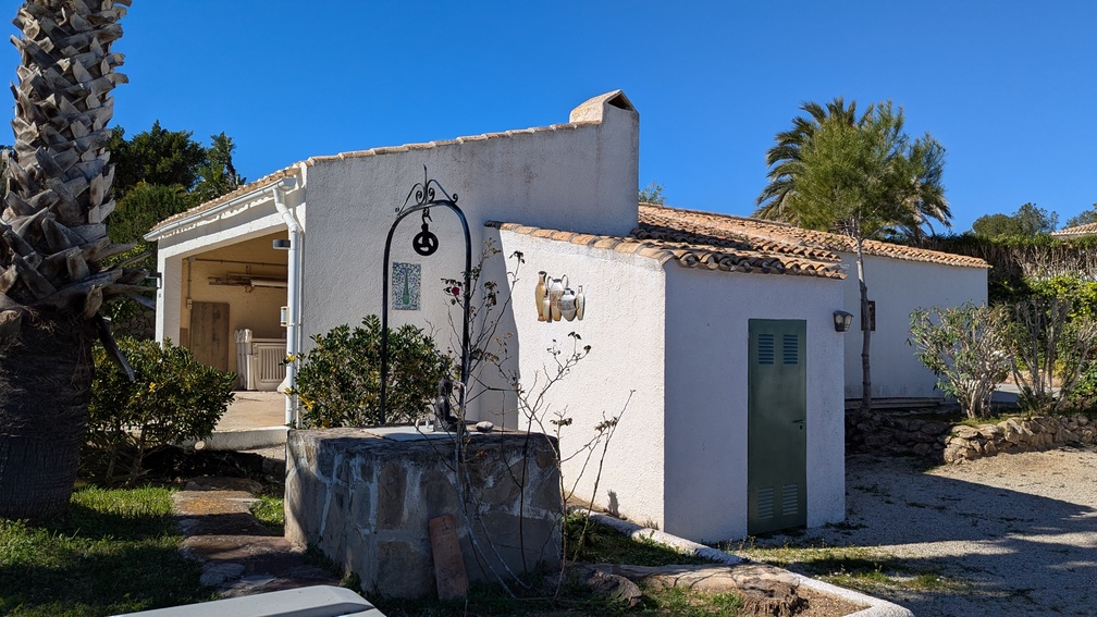 Exterior de una construcción anexa tradicional con fachada blanca, tejado de teja cerámica y un pozo decorativo de piedra con arco de hierro forjado.