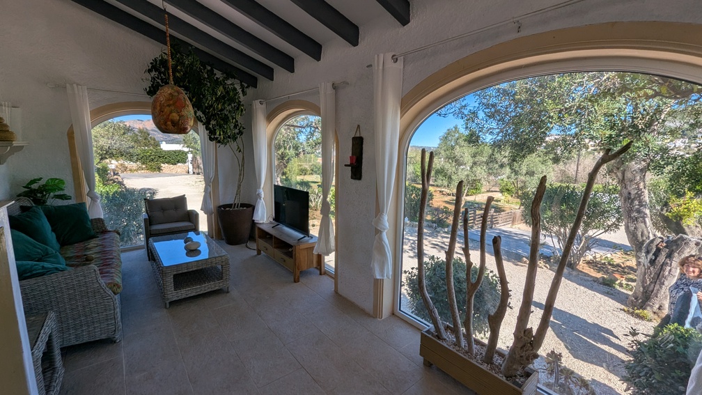 Naya acristalada con vigas de madera oscura, amplios ventanales en arco con vistas al campo y zona de estar con muebles de mimbre y suelo cerámico.